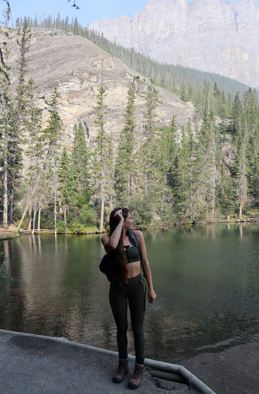 A picture containing a hiker looking behind them standing outdoors in front of Grassi Lake that has clear blue and green water reflecting the pine trees above backed by a mountain landscape and clear blue sky