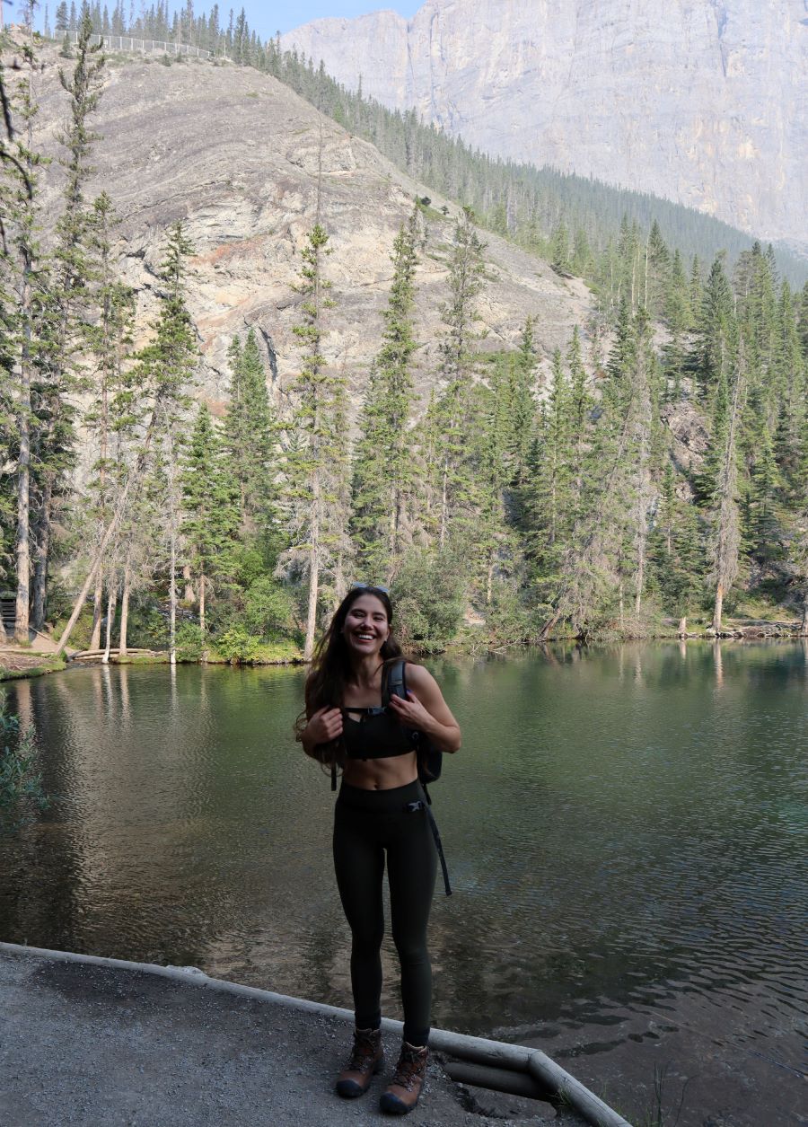 A picture containing a hiker laughing at the photographer and standing outdoors in front of Grassi Lake that has clear blue and green water reflecting the pine trees above backed by a mountain landscape and clear blue sky