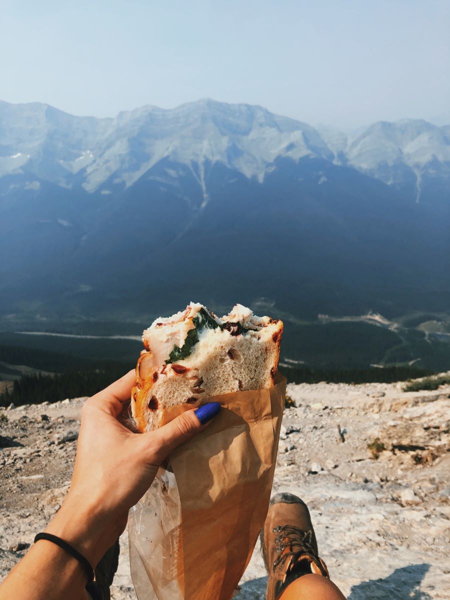 manicured hands with blue nails of a hiker wearing brown hiking boots holding a half-eaten sandwich in front of a hazy Mount Lady Macdonald along Ha Ling Peak's saddle ridge