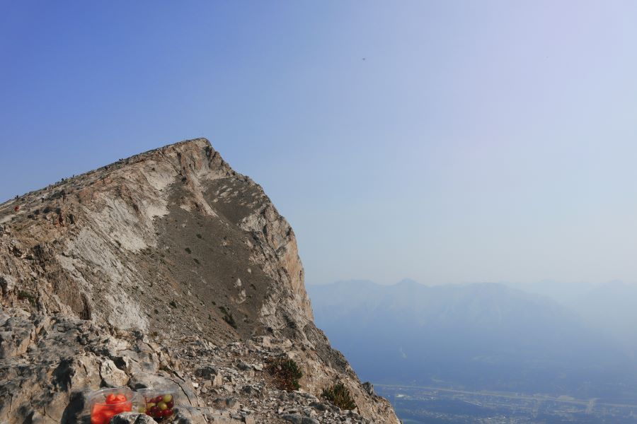 two plastic containers of watermelong and grapes pictured in front of the summit of Ha Ling Peak overlooking a hazy Canmore, Alberta, Canada, and also pictured are numerous hikers scrambling up Ha Ling Peak's summit