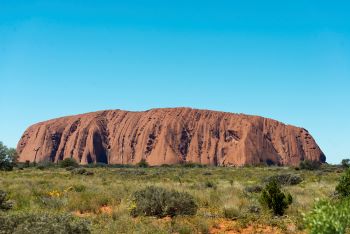 image of famous landmark called Uluru centred in front of green and red dusty landscape on a bright, sunny, bluebird day